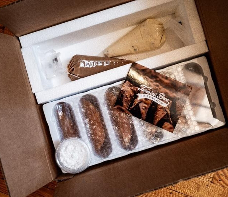Hand holding a filled cannoli above a tray of empty cannoli shells lined with bubble wrap.